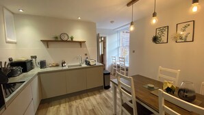 A modern kitchen with a central island, white cabinetry, and a dining area with a wooden table and white chairs. -  Coachman’s Cottage at Brylach Steading, Speyside (Aberlour)