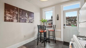 Un coin repas avec une table et deux chaises, un tableau représentant un paysage urbain, une fenêtre donnant sur la vue et un réfrigérateur.