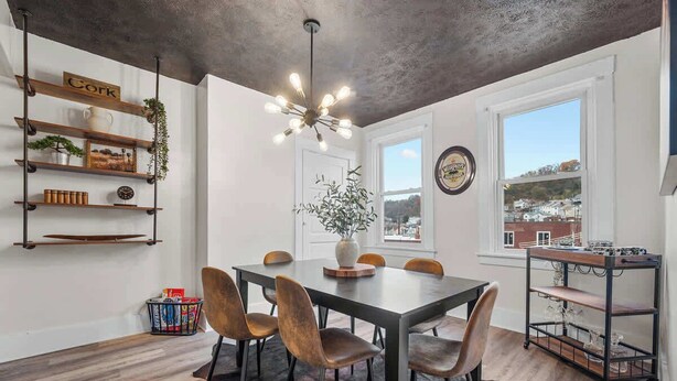 Un coin repas comprenant une grande table, six chaises, un luminaire suspendu, une étagère avec divers objets et une fenêtre donnant sur des bâtiments.