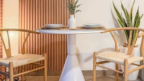Un área de comedor con una mesa blanca, dos sillas, un florero con una planta y una planta alta junto a la mesa.