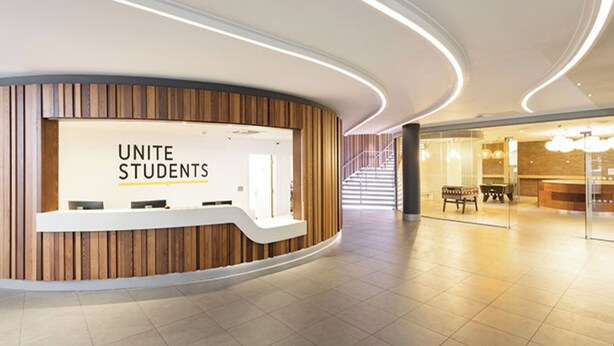 Un área de recepción moderna con un cartel de 'UNITE STUDENTS', un mostrador de recepción de madera y una escalera que conduce a un nivel superior.