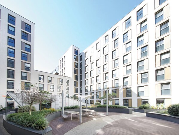 A modern building with multiple floors, large windows, and a courtyard with seating areas and greenery. - Student Only Stylish Ensuite at Drapery Place near Tower Hamlets (London)