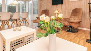 A room with a white table, a vase with flowers, brown chairs, and a television on the wall.