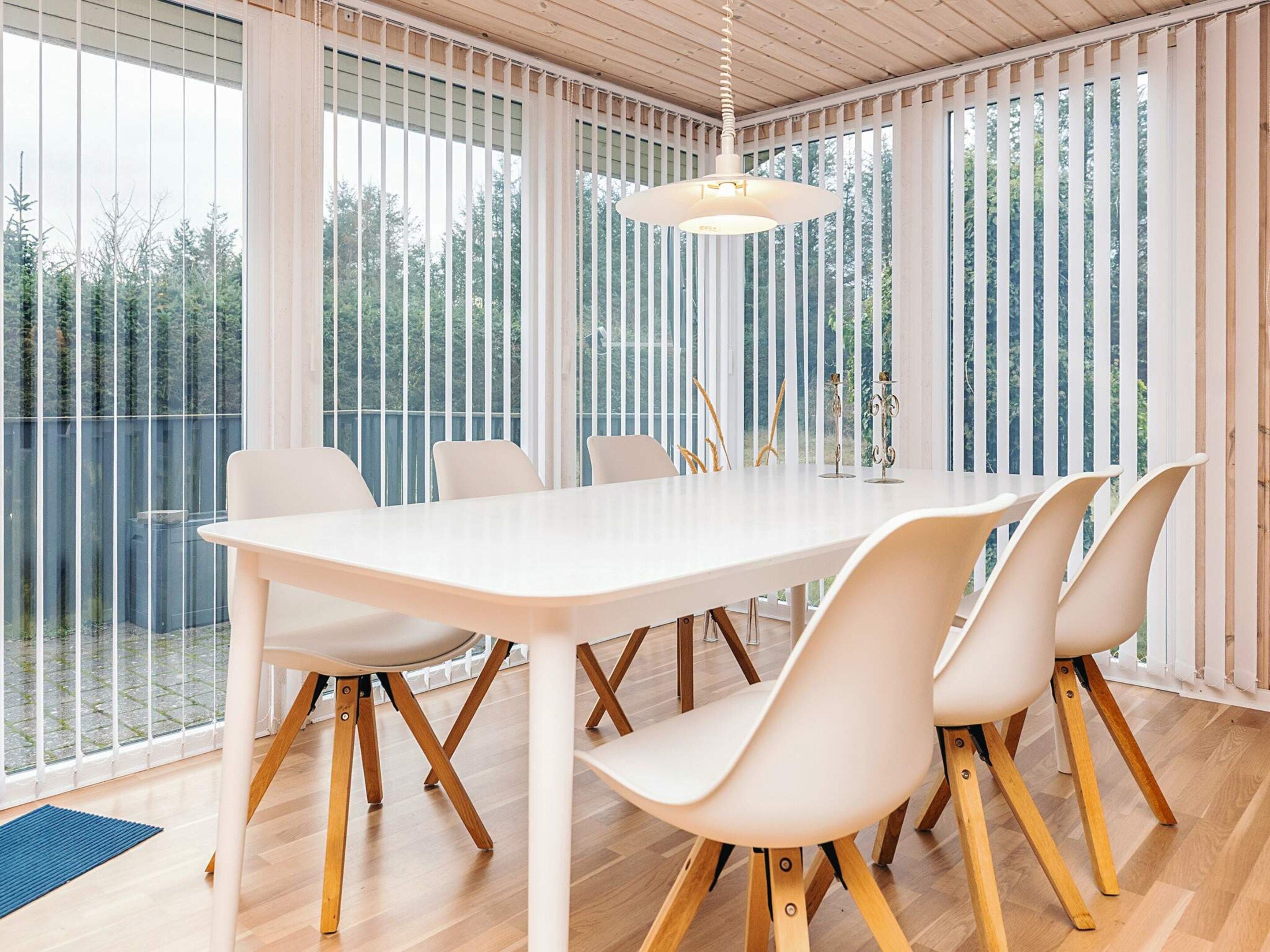 Un comedor moderno con una mesa y sillas blancas, ventanas amplias con persianas, y una lámpara colgante.