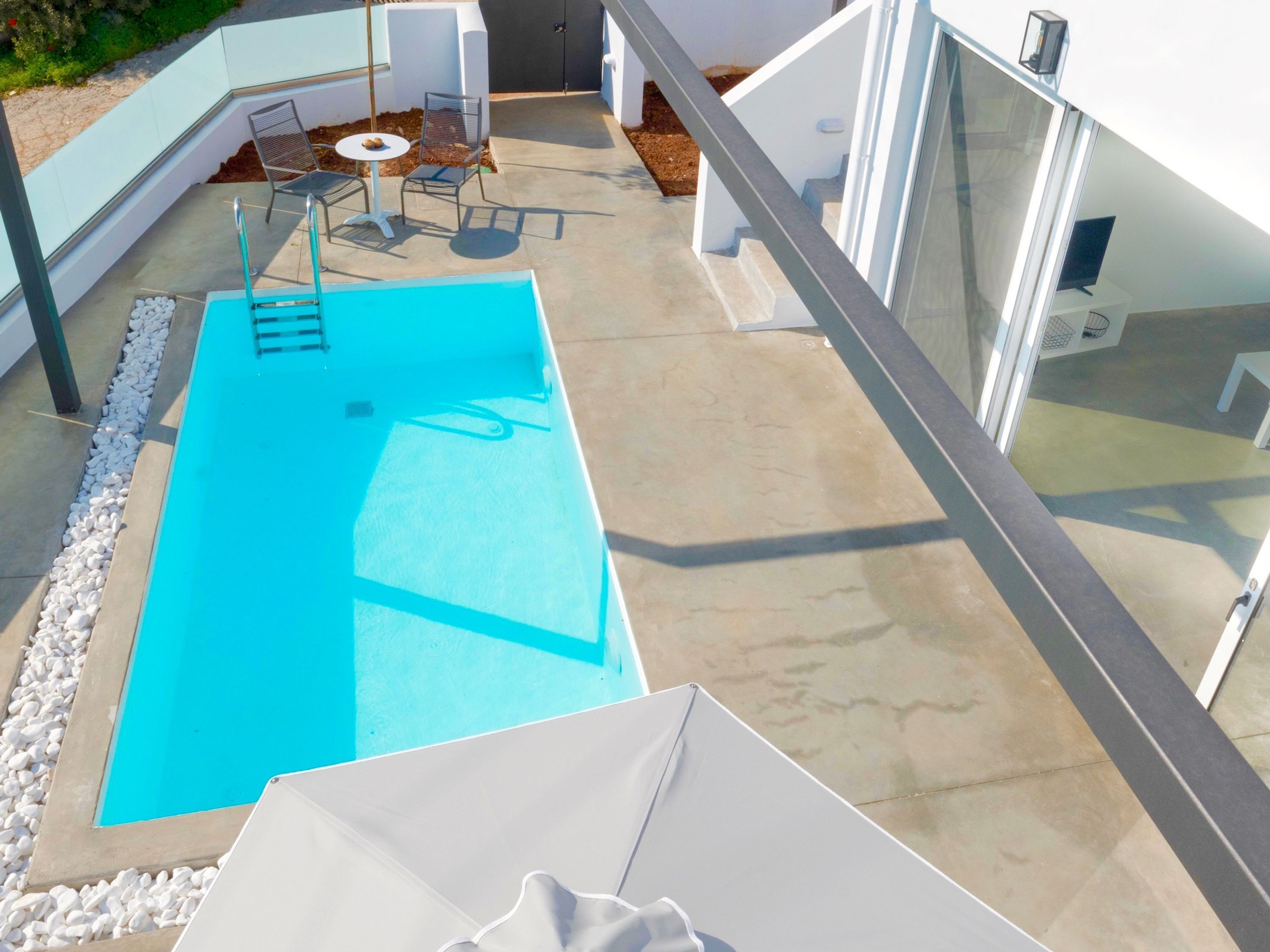 A modern outdoor swimming pool with a clear blue pool, surrounded by white pebbles. There are metal chairs and a small round table with a white umbrella.