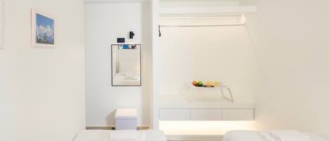 Habitación de hotel minimalista con dos camas, una mesita de noche pequeña, un espejo y una estantería con artículos de aseo.