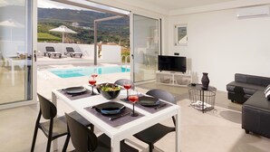 A modern living room with a pool view, a dining area with a table set for two, and a sofa. - Villa Paradiso 2 With Private Pool (Malevizi)