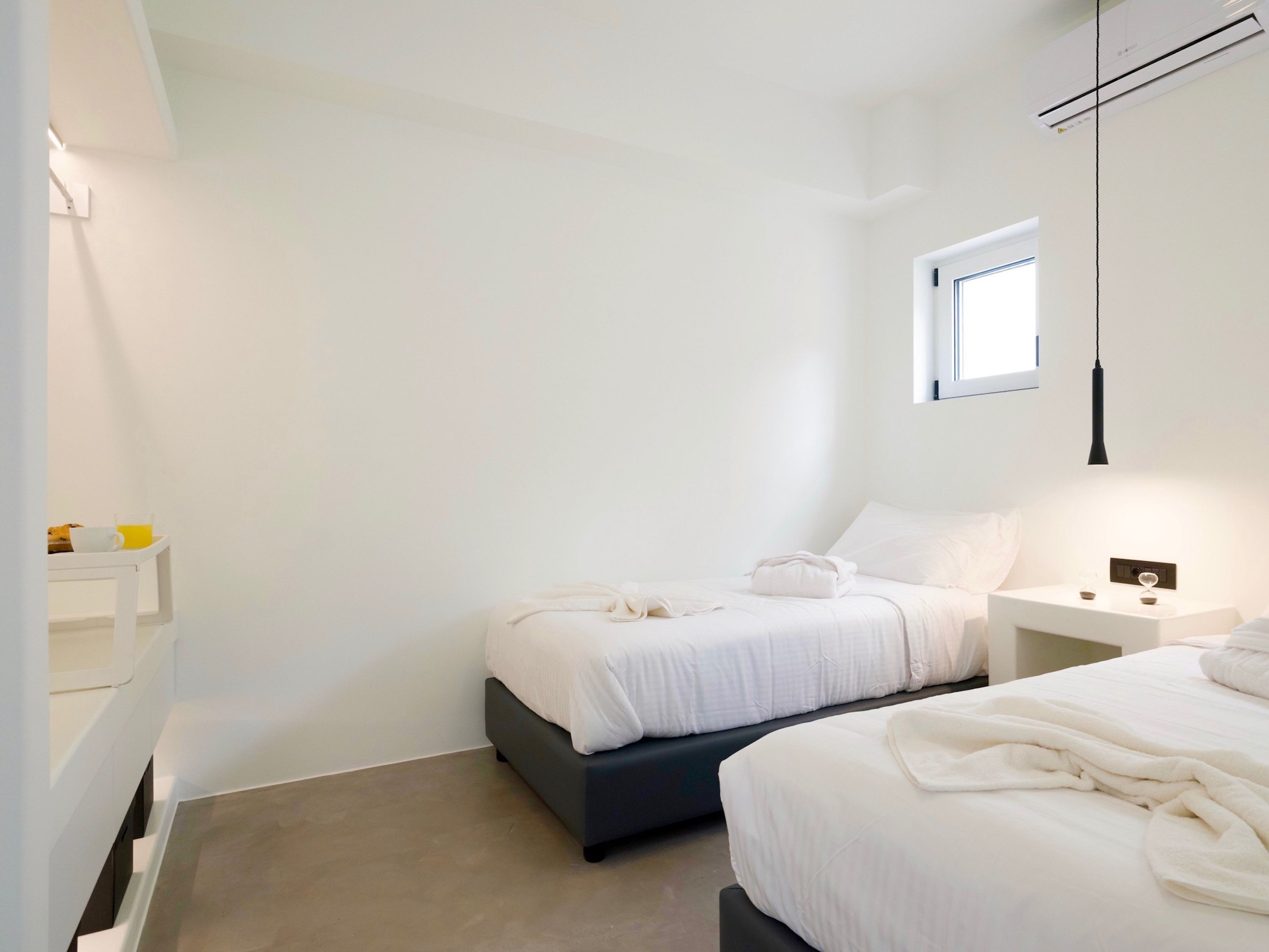 A minimalist bedroom with two beds, a small bedside table, a nightstand, and a window.