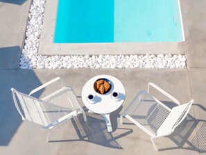 Un área junto a la piscina con dos sillas de metal blanco, una mesa redonda con un bol de comida y bebidas, y una piscina con borde de guijarros.