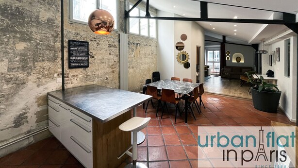 A modern kitchen with a marble countertop, wooden cabinets, and a dining area with a round table and chairs. There is a large window providing natural light and a view of the outside.
