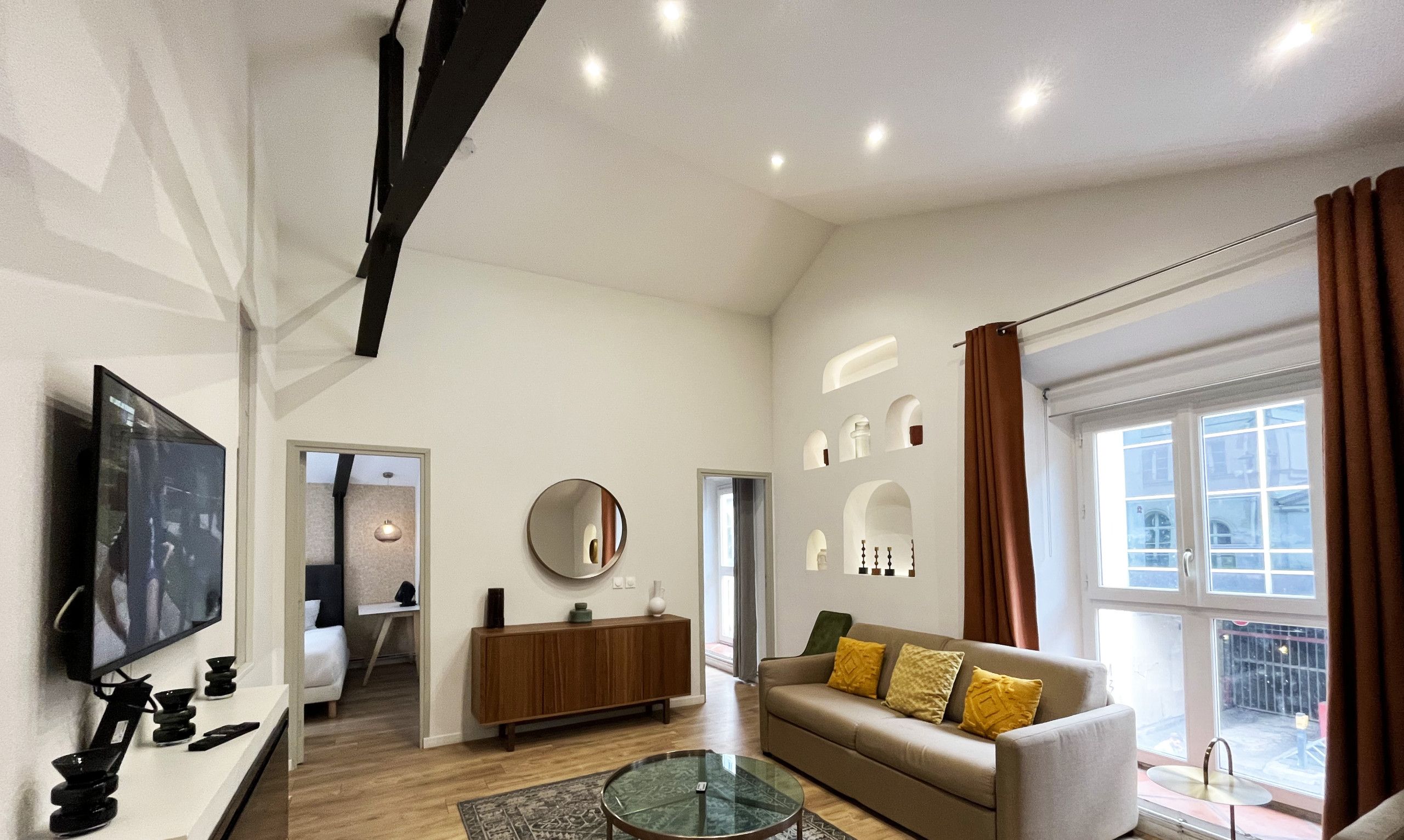 A modern living room with a sofa, coffee table, and TV. There is a window with curtains, a mirror, and a small cabinet.