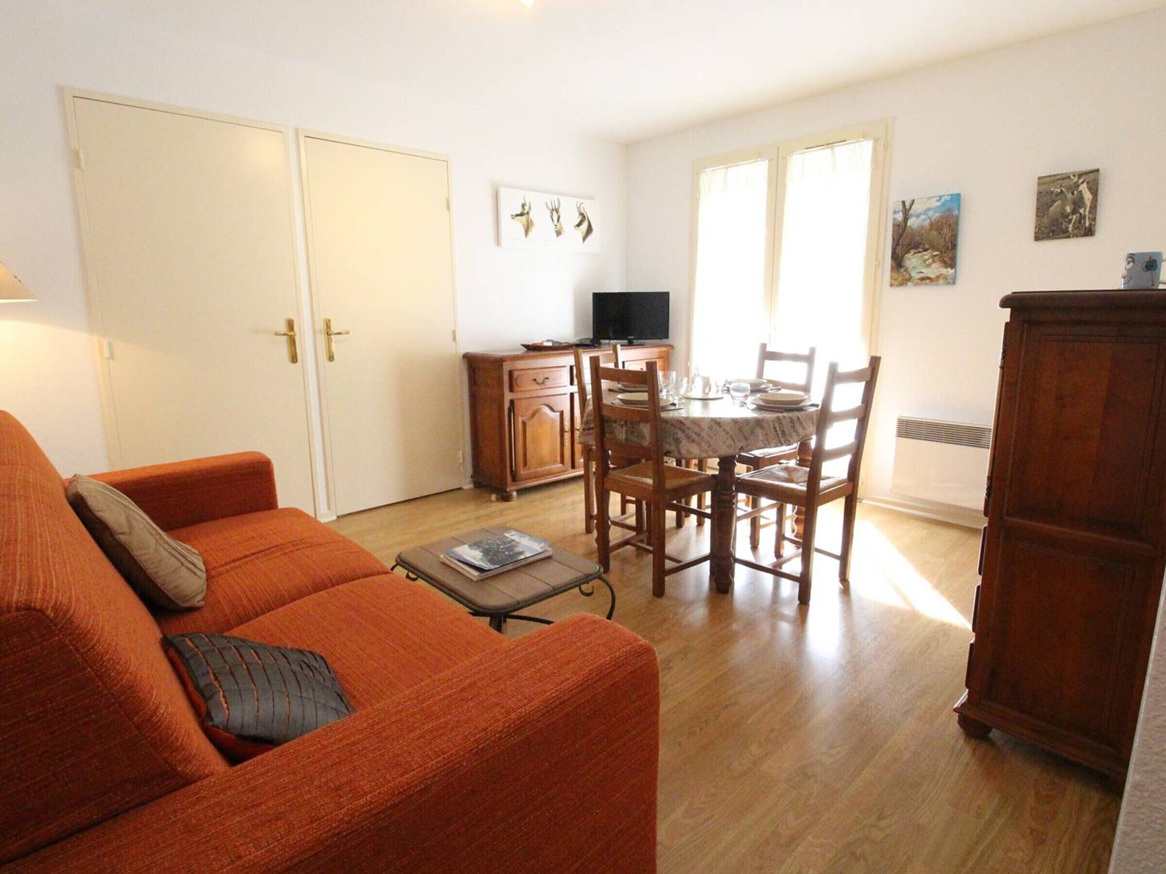 A living room with an orange sofa, a dining table with chairs, a TV, and artwork on the wall.