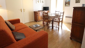 A living room with an orange sofa, a dining table with chairs, a TV, and artwork on the wall. - T2 Luchon Center - Balcony - Parking Included - Pets Welcome (Bagnères-de-Luchon)