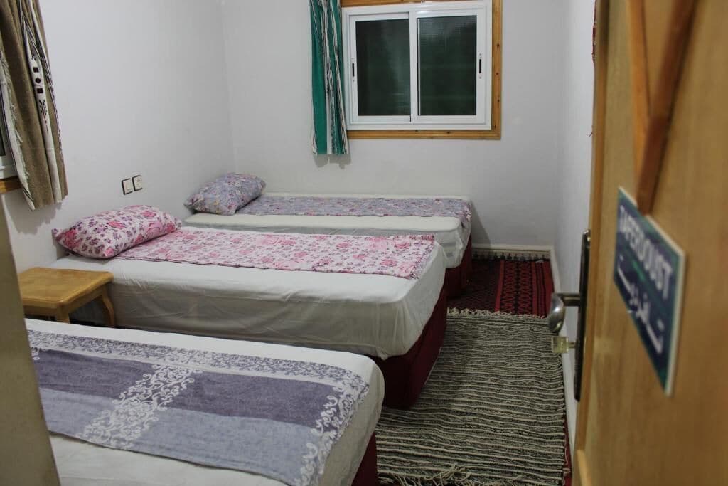 A small, clean room with three beds, a window with curtains, a wooden door, and a rug on the floor.