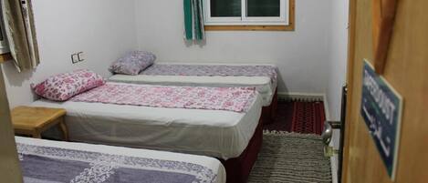 A small, clean room with three beds, a window with curtains, a wooden door, and a rug on the floor.