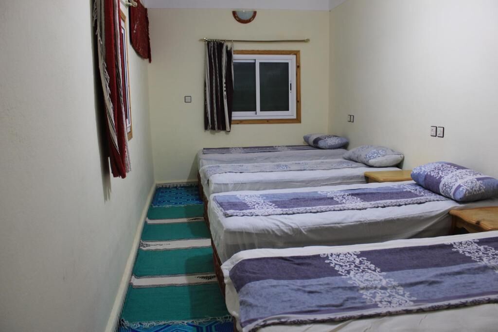 A room with three beds, each with a patterned bedspread and pillows. There is a window with curtains, a wall-mounted light fixture, and a small wooden stool.