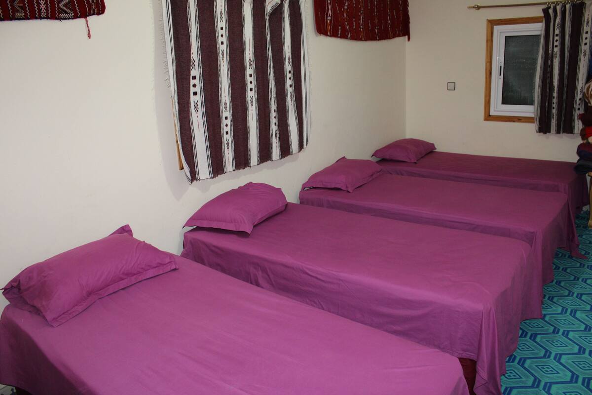 Une chambre avec trois lits, chacun recouvert d’une couvre-lit violette et agrémenté d’oreillers assortis. Il y a une fenêtre avec des rideaux, et une décoration murale à motifs au-dessus des lits.