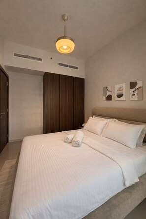 Una cama bien tendida con sábanas blancas y dos toallas enrolladas, un armario de madera y una lámpara en el techo.