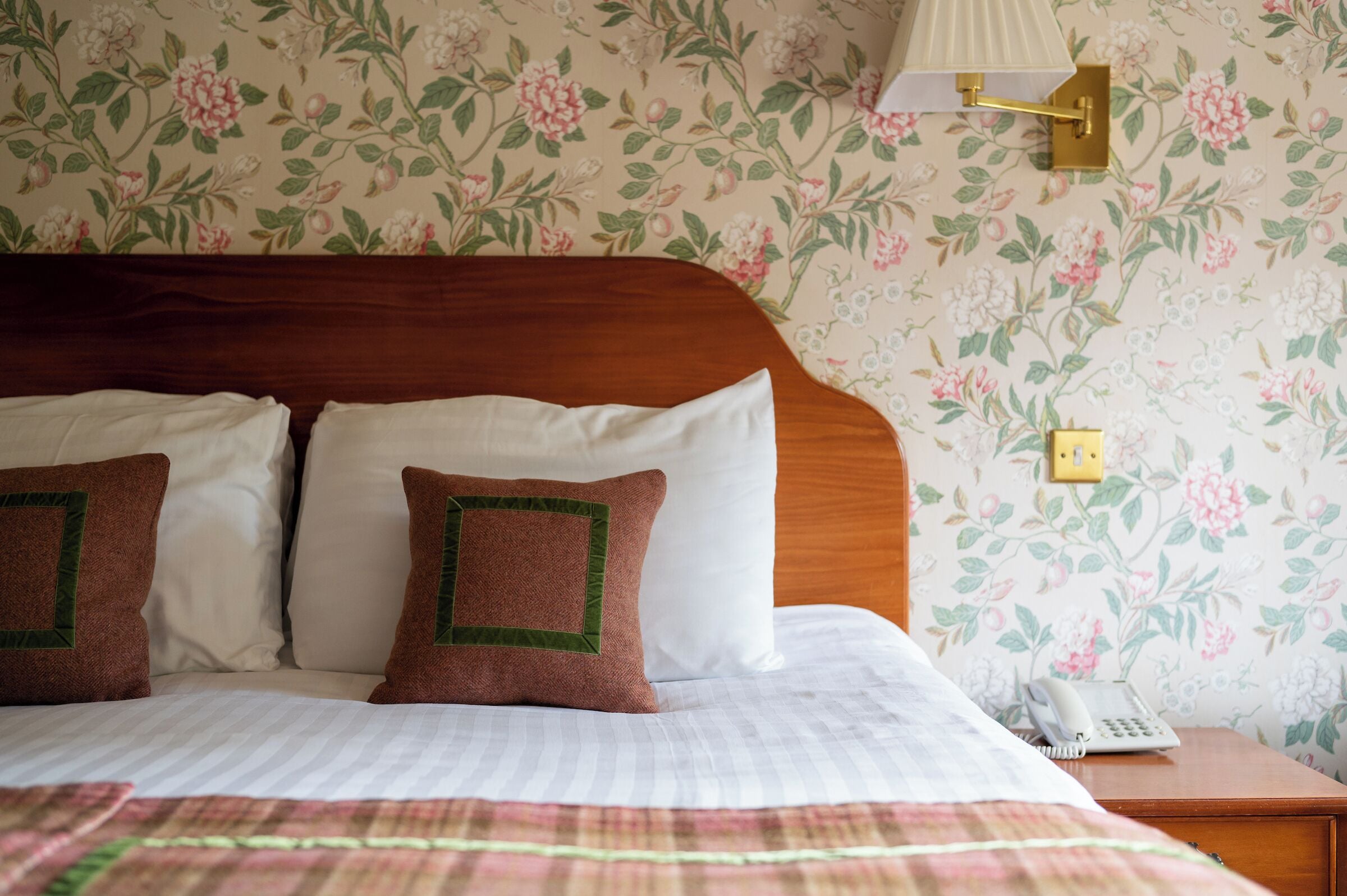 A neatly made bed with a wooden headboard, white and brown pillows, and a floral wallpaper pattern.