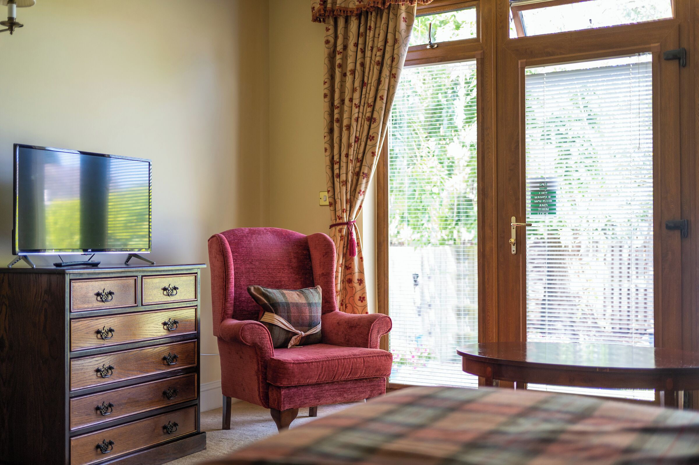 Une pièce comprenant une commode en bois, un fauteuil rouge avec un coussin à carreaux et une porte-fenêtre coulissante avec des stores.
