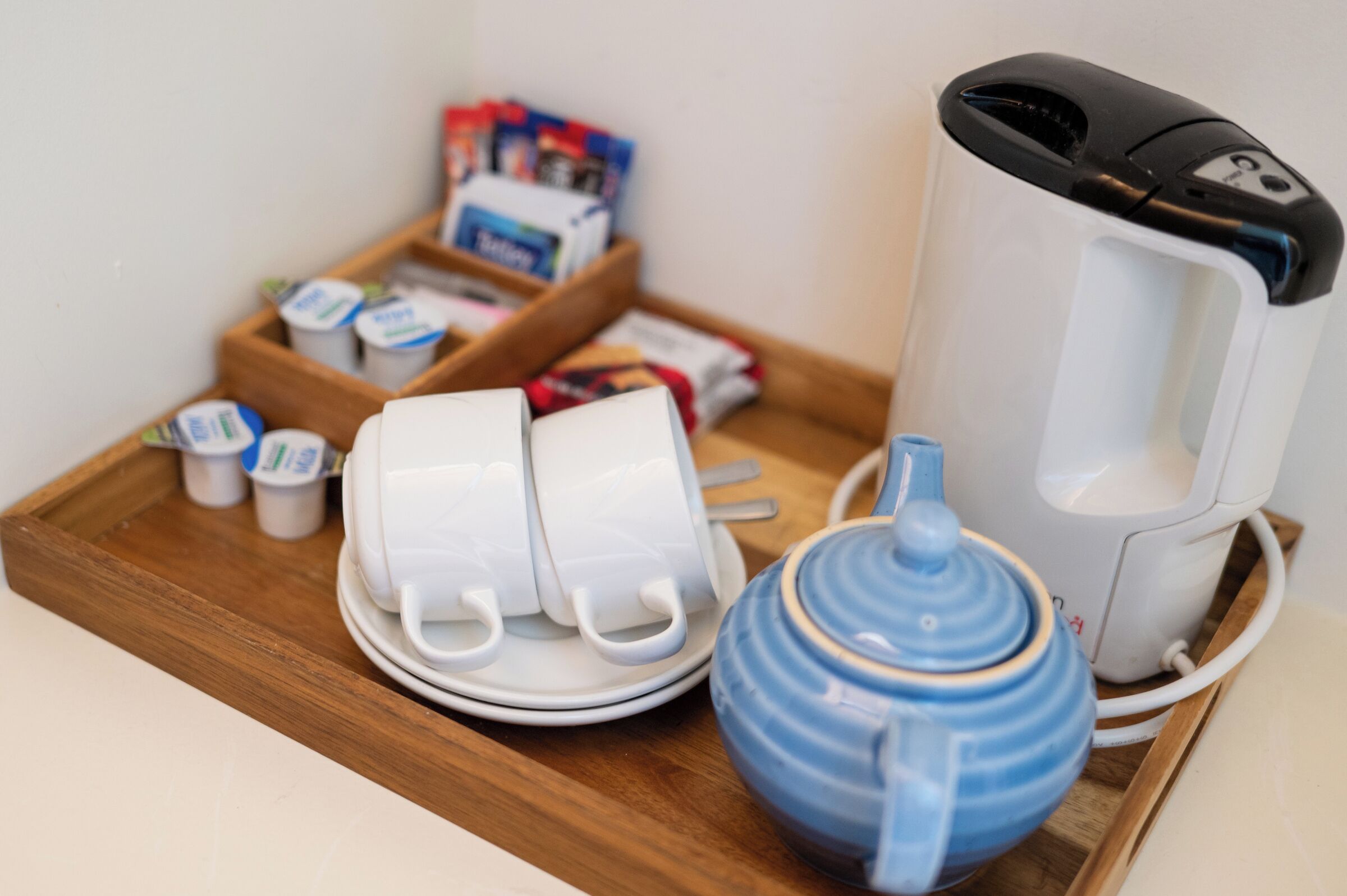 A tray with a white electric kettle, a blue teapot, two white cups, and a set of small containers on a wooden tray.