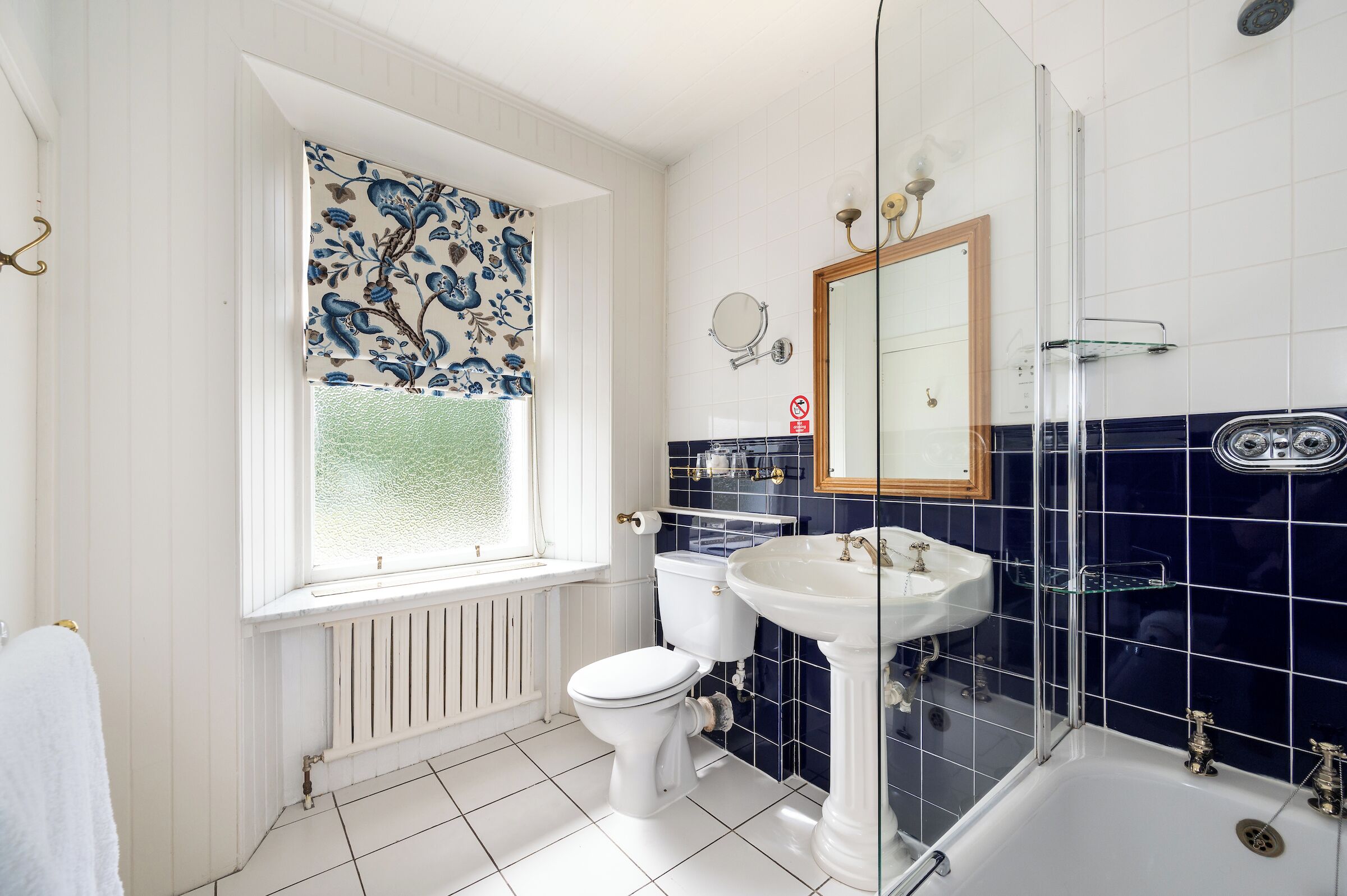 A bathroom with a white toilet, a white sink, a glass shower enclosure, and a window with patterned curtains.
