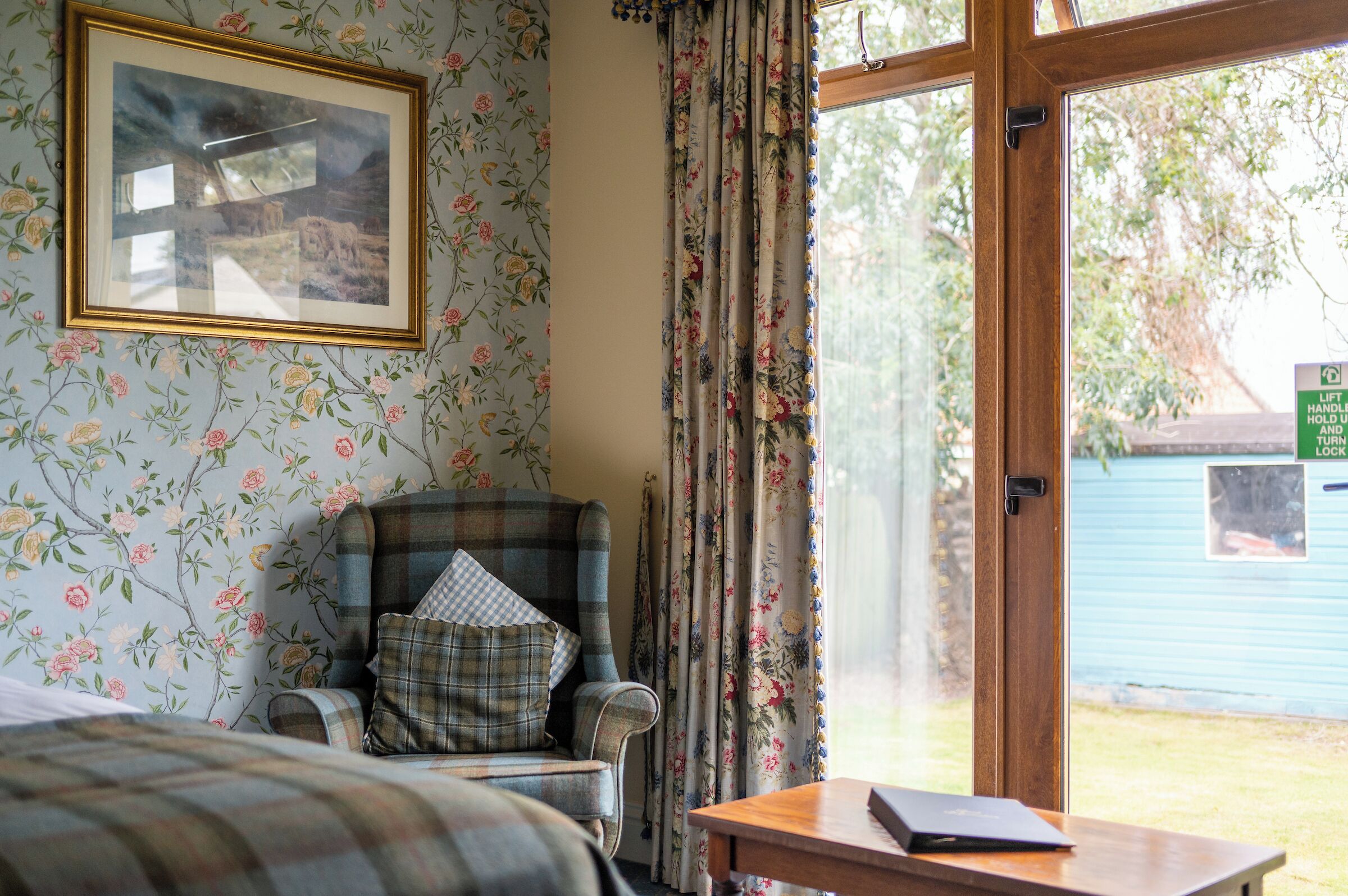 A room with a plaid armchair, a bed, a wooden table, and a window with curtains.
