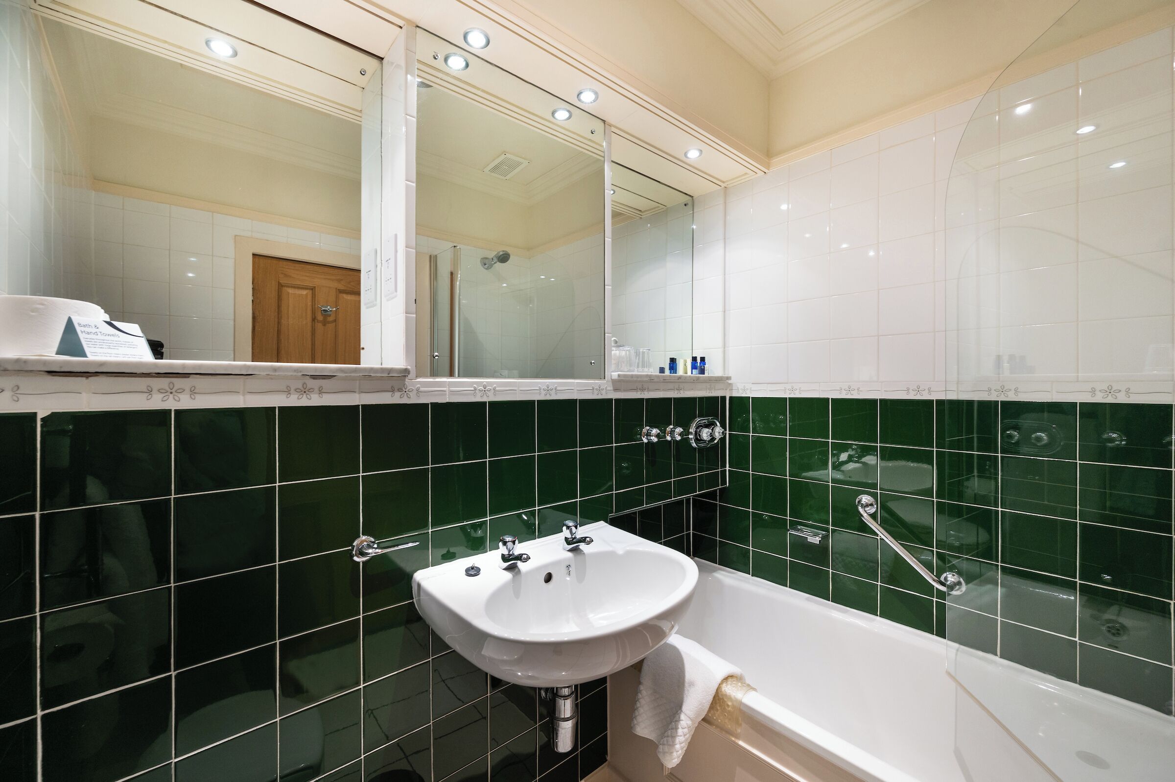 Un baño con lavamanos blanco, paredes con azulejos verdes, un espejo grande y una bañera.