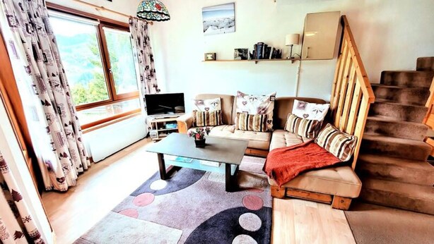 Una sala de estar acogedora con un sofá, una mesa de centro y un televisor. Se observa una escalera a la derecha y una ventana con cortinas.