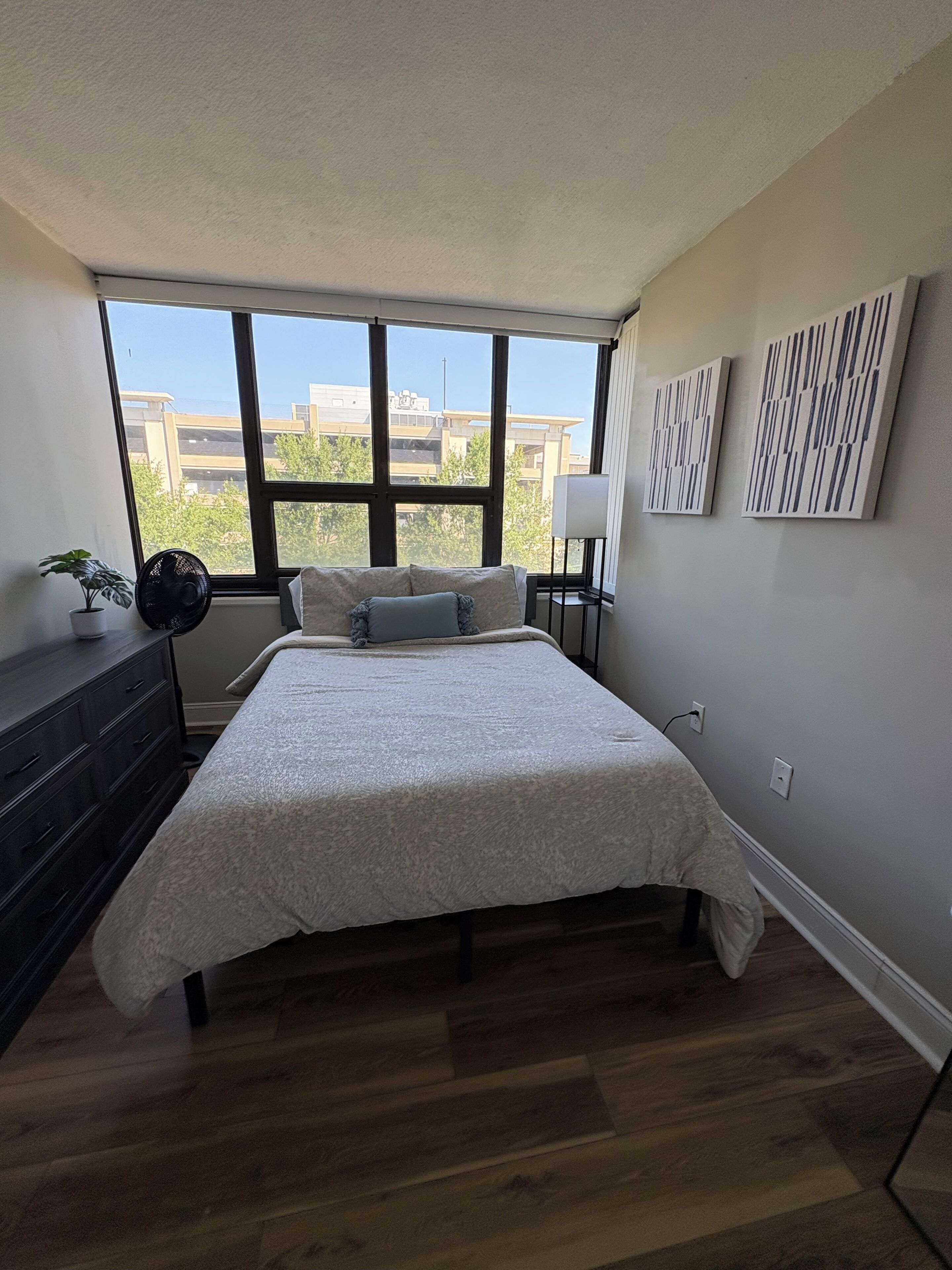 A bedroom with a large window, a bed with a white bedspread, a dresser, a plant, and two wall-mounted lamps.
