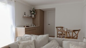 A modern living room with a sofa, a dining table with chairs, a kitchen area with wooden cabinets, and a white door. - Costal Comfort (Nelson Bay)