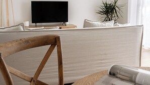 A modern living room with a wooden chair, a sofa, a flat-screen TV, a plant, and a magazine on a wooden table. - Costal Comfort (Nelson Bay)