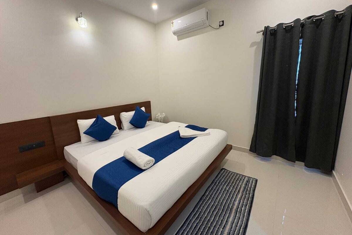 A hotel room with a large bed, a wooden headboard, a bedside table, a wall-mounted air conditioner, and a window with black curtains.