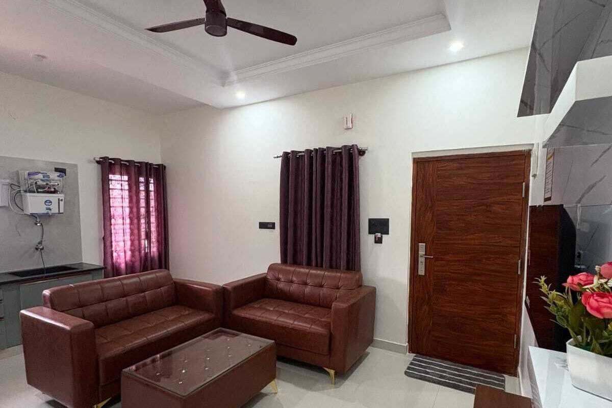 A living room with a sectional sofa, a coffee table, a ceiling fan, a wooden door, and a kitchen area with a water heater.