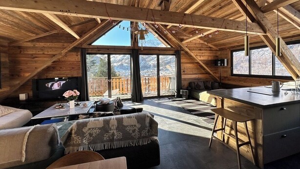 A cozy attic room with a wooden ceiling, a large window, a sofa, a coffee table, and a kitchen area with a bar stool. - CHALET LE CAIRN view of the village Large open spaces 5 minutes from the cable car (Valloire)