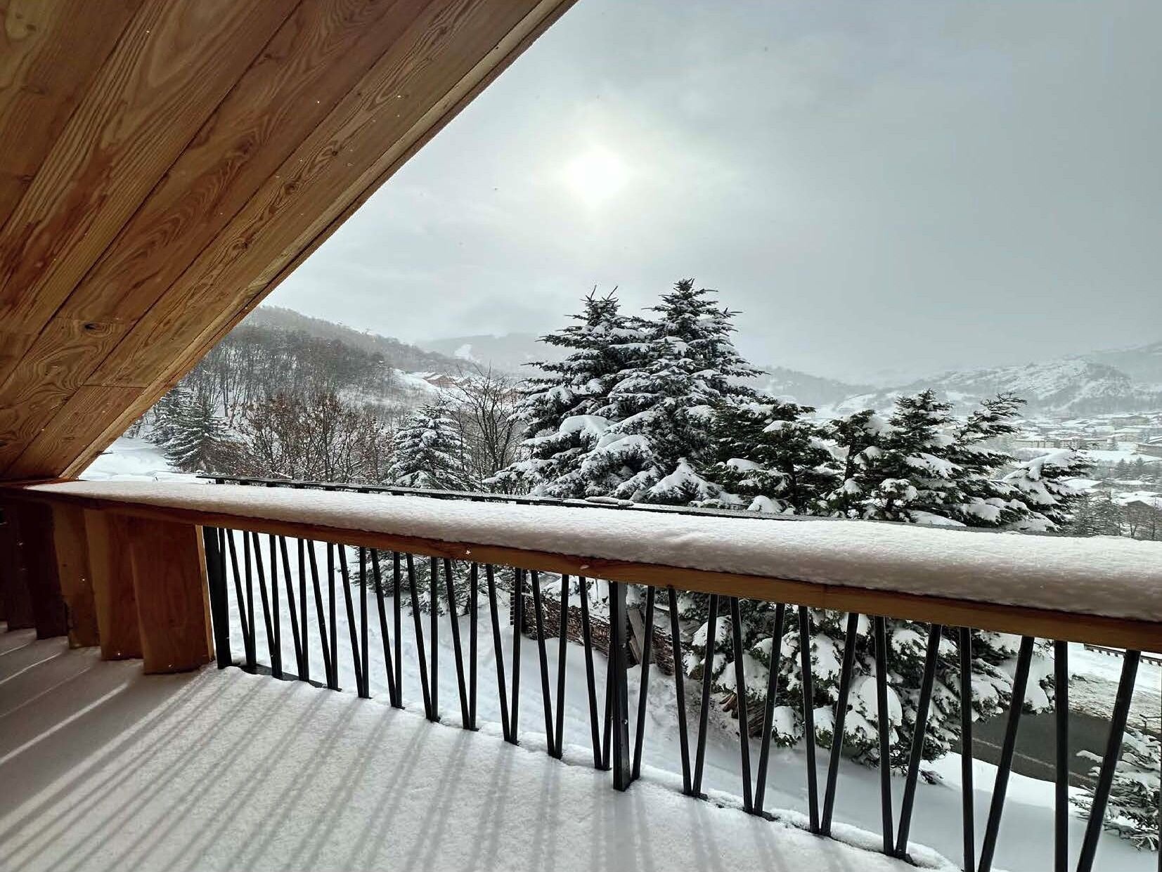 Ein Holzbalkon mit Metallgeländer, der auf eine verschneite Landschaft mit Bäumen und Bergen blickt.