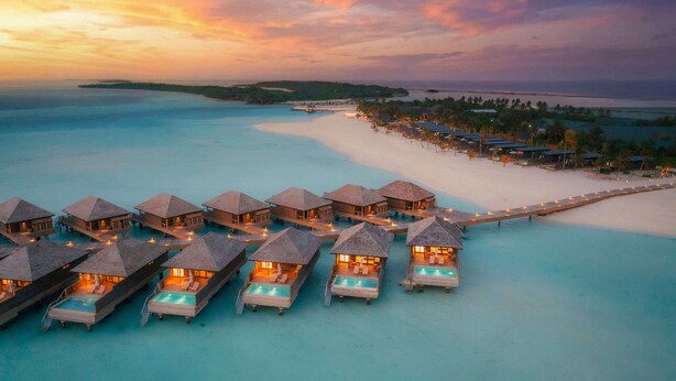 Bungalows sobre el agua con piscinas infinitas, unidos por una pasarela, con vistas a una playa y al océano.