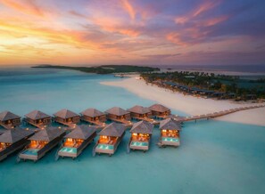 Des bungalows sur pilotis dotés de piscines à débordement, reliés par une jetée, et offrant une vue sur une plage et l’océan.