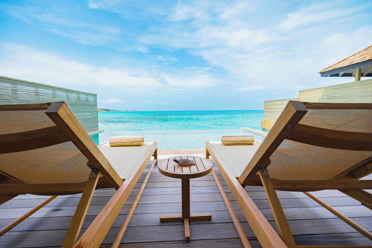 Een houten terras met twee loungestoelen en een kleine tafel, met uitzicht op een heldere blauwe zee en een zwembad.