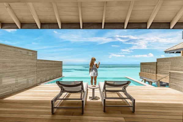 Une terrasse en bois avec deux chaises longues et une petite table, offrant une vue sur un océan d’un bleu limpide et une structure en bois construite sur pilotis au-dessus de l’eau.