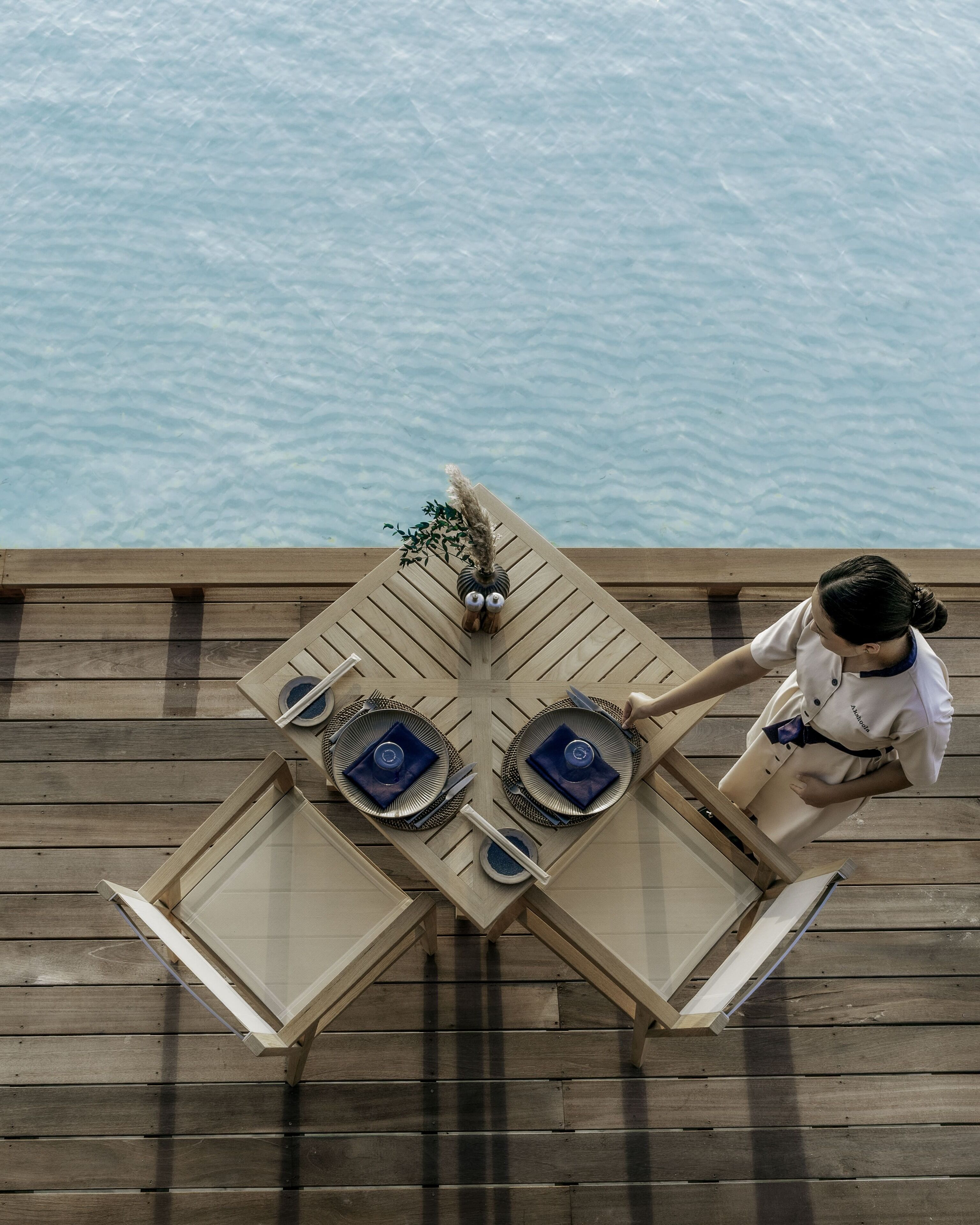 A dining table with a decorative owl centerpiece, set with blue napkins and silverware, on a wooden deck by a body of water.