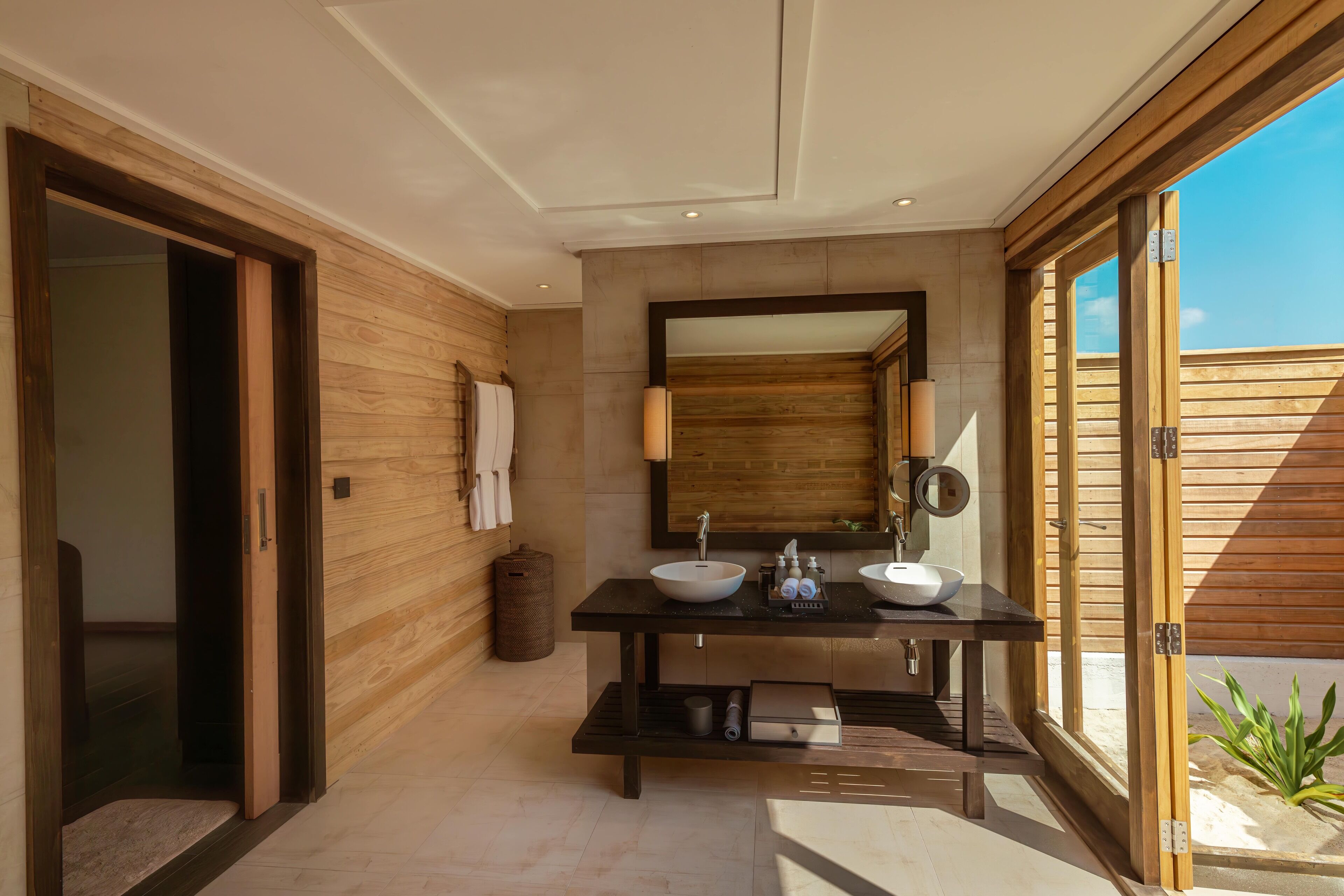 A modern bathroom with a wooden interior, a large mirror, two sinks, and a view of the outdoors.