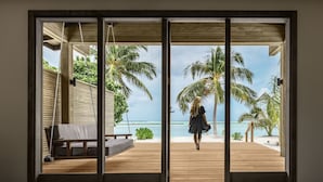 A room with a large sliding glass door leading to a wooden deck overlooking a beach with palm trees and the ocean. - Nala Maldives by Jawakara (Innahuraa)
