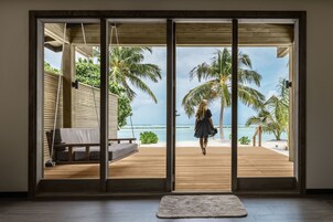 Habitación con un amplio ventanal corredero que da a una terraza de madera con vista a una playa con palmeras y el océano.