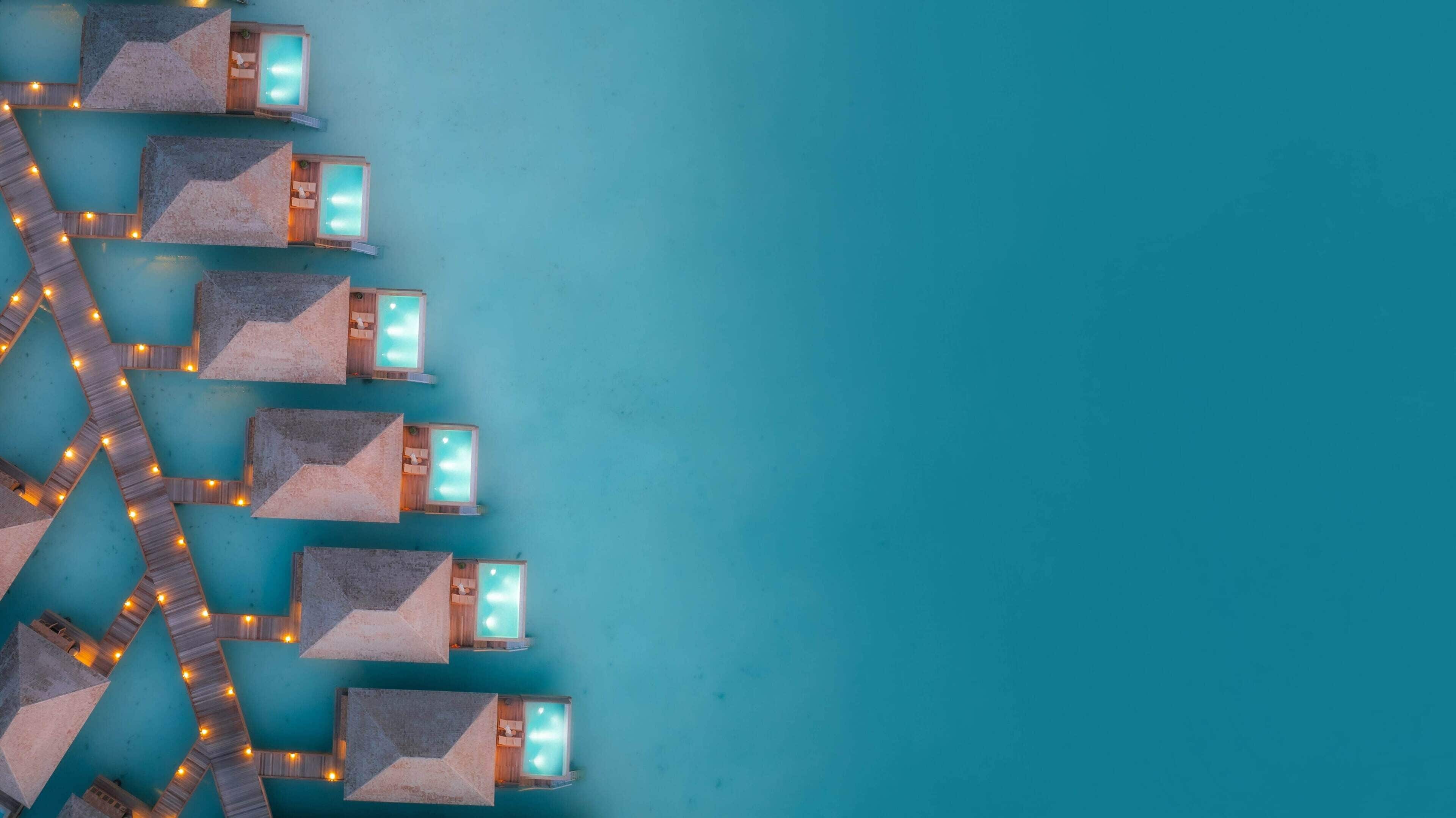 An aerial view of a resort with multiple rectangular pools, each with a wooden deck and a small seating area, connected by a wooden pathway with lights.