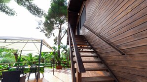 Un escalier en bois mène à un espace extérieur abrité, aménagé avec des tables et des chaises, et entouré de verdure.