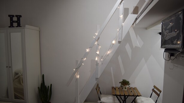 Uma escadaria decorada com luzes de fada, uma mesa de jantar de madeira com cadeiras, uma planta em vaso e um armário branco com espelho.
