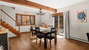 A dining area with a wooden table and chairs, a chandelier, and a view of the outdoors through a large window. - Snow Haus | Hot Tub & Bar | 10 Min to Mount Snow! (Wardsboro)