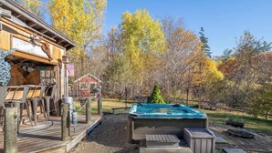 A wooden deck with outdoor furniture, a hot tub, and a rustic building with a sign. - Snow Haus | Hot Tub & Bar | 10 Min to Mount Snow! (Wardsboro)