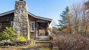 A rustic log cabin with a stone chimney, a porch, and a forested backdrop. - Snow Haus | Hot Tub & Bar | 10 Min to Mount Snow! (Wardsboro)